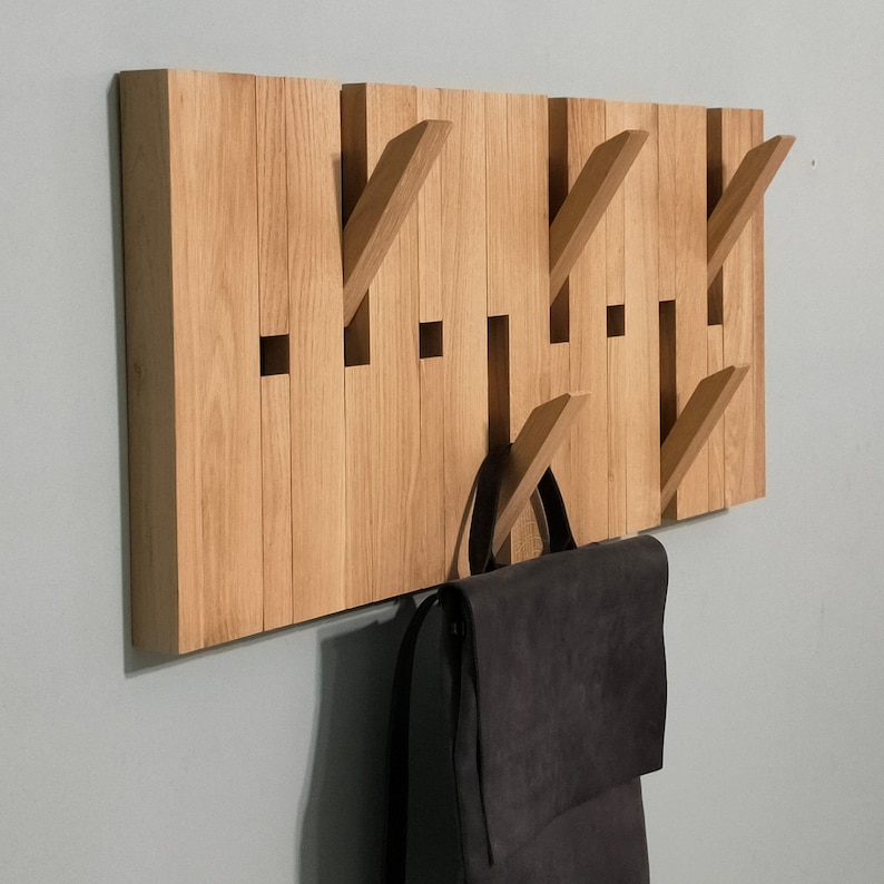 May include: A wooden wall-mounted coat rack with multiple hooks. The rack is made of light brown wood and has a natural finish. A black backpack is hanging on one of the hooks.