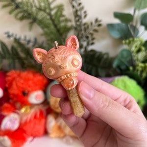 May include: A pink and orange ceramic cat charm with a crystal pendant. The charm is attached to a brown crystal point.
