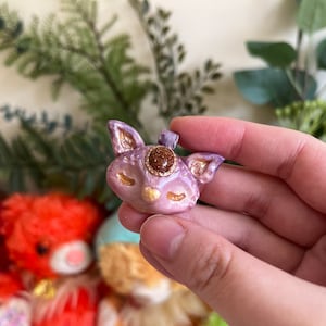 May include: A purple and gold clay pendant shaped like a fox with a closed eye and a gemstone in the center of its forehead.