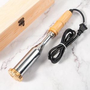 Könnte beinhalten: Eine Holzkiste mit einem Metall-Logo und einem elektrischen Branding-Eisen aus Silber und Holz mit einem schwarzen Kabel und Stecker.