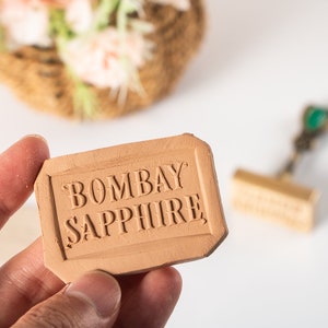 May include: A rectangular, light brown stamp with the words "BOMBAY SAPPHIRE" embossed on it. The stamp is held in a hand, with a blurred background featuring a basket of flowers and a gold-colored stamp.