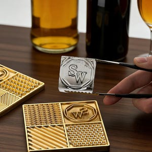 Könnte beinhalten: Ein quadratischer Eisstempel mit den Buchstaben "SW", der mit einer Zange gehalten wird. Der Stempel ist silberfarben und befindet sich über einem goldfarbenen Tablett mit verschiedenen strukturierten Designs. Flaschen und ein Glas sind im Hintergrund.