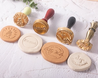 May include: A set of six gold-toned metal stamps with wooden handles. The stamps feature various designs, including a crown, a flame, a floral wreath, and text. The stamps are used to create impressions on clay or wax.