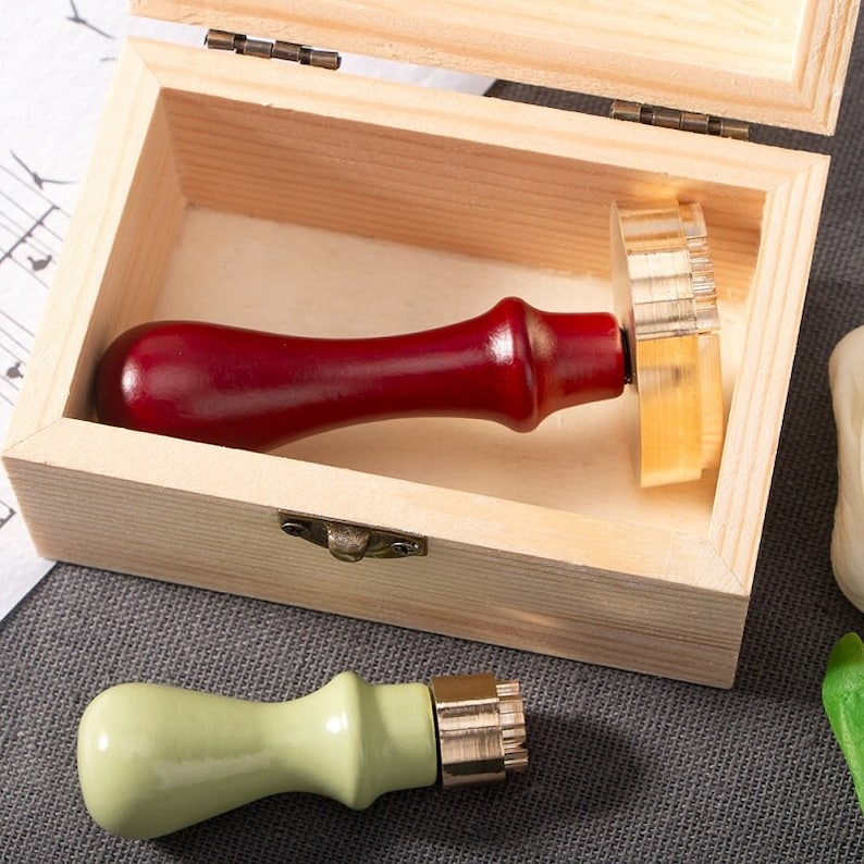 May include: A wooden box with a hinged lid contains a red-handled wax stamp. A second stamp with a light green handle rests outside the box. The stamps have a decorative design on the stamp head.