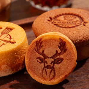 Puede incluir: Tres productos horneados redondos, de color marrón dorado, con diferentes diseños. Uno tiene una cabeza de ciervo, otro tiene un logotipo con el texto "Le Thomas" y el tercero tiene un diseño de un pájaro estilizado.