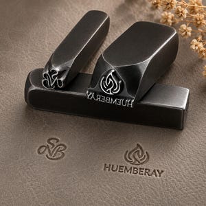 May include: Two dark metal stamping tools with rectangular shafts. One tool features a stylised monogram of the letters SNB, while the other displays a flame-like emblem above the text HUEMBERAY. Both stamps leave clear impressions on the surface.