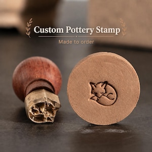 May include: A custom pottery stamp with a wooden handle and a brass head, accompanied by a circular clay impression featuring a debossed design of a curled-up sleeping fox. The text Custom Pottery Stamp Made to order is visible above.