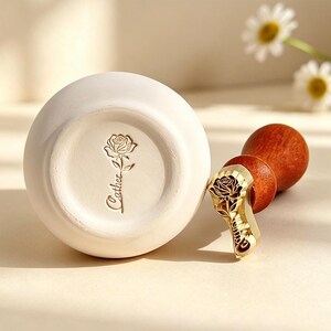 May include: A white ceramic vase with a rose design and the word "Gather" engraved on the bottom. A gold-colored stamp with a rose design and a wooden handle is next to the vase.
