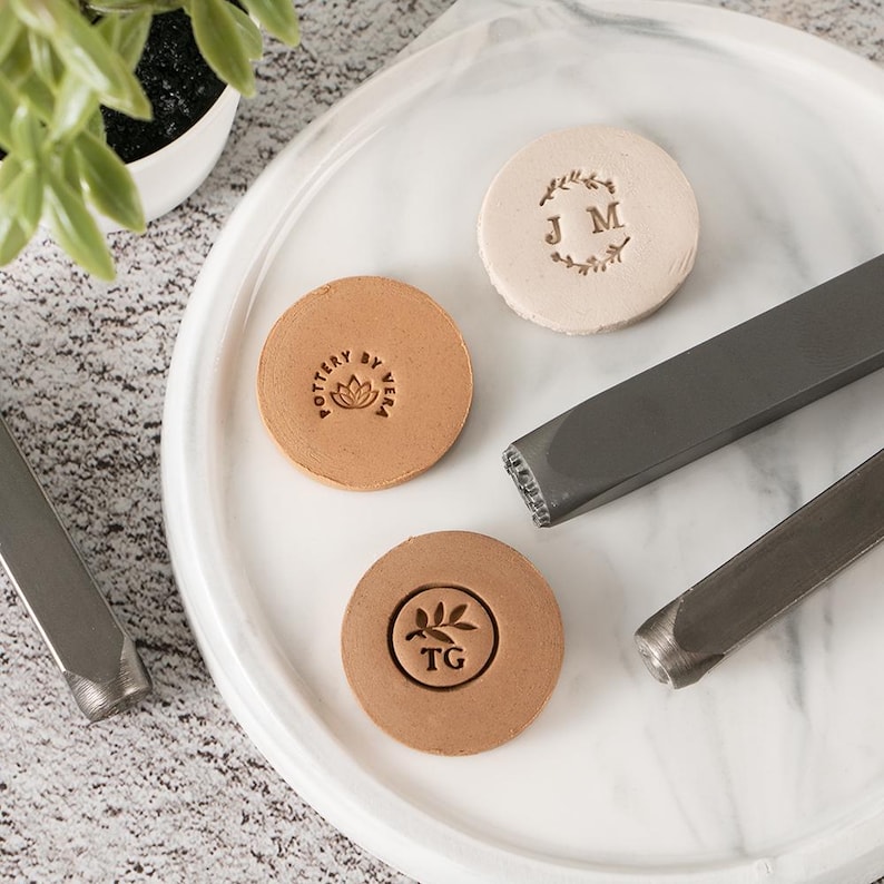 May include: Three circular clay discs with stamped designs and two metal stamping tools. One disc is light beige with the letters JM inside a leaf wreath. Two tan discs feature a lotus flower with the text POTTERY BY VERA and the letters TG.