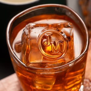 Könnte beinhalten: Nahaufnahme eines Glases mit bernsteinfarbener Flüssigkeit und einem großen Eiswürfel. Der Eiswürfel trägt die Initialen "JM" in einem kreisförmigen Design. Das Glas ist klar und hat einen dicken Boden.
