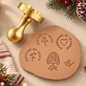 May include: A gold-colored metal pottery stamp with a smooth handle and a flat circular base. Next to it, a tan clay disc features four distinct embossed designs: a flower with text, a heart with text, a flower with text, and a dog with text.