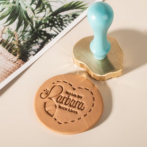 May include: A round, brown leather stamp with the text "Made by Barbara with love" in a cursive font. The stamp is surrounded by a dashed line. A blue wooden handle and a gold metal base are also visible.