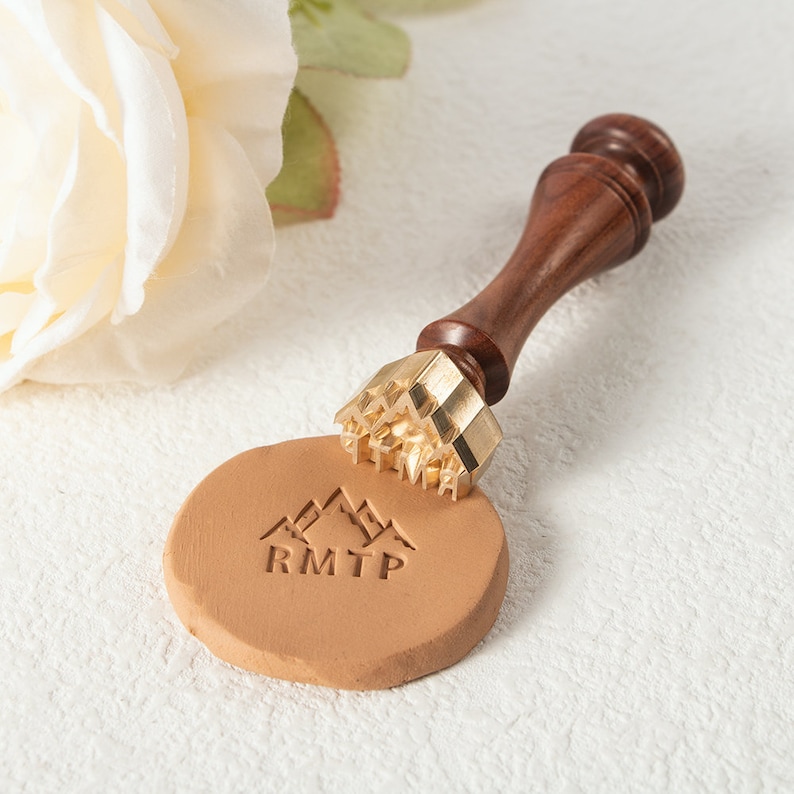 May include: A brass wax seal stamp with a wooden handle is pressed into a tan wax seal. The stamp creates a mountain and text design. The handle is dark brown, and the wax seal is round. A white rose is in the background.