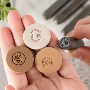 May include: Three circular clay discs with stamped designs and a metal pottery stamp. One disc is off-white with a wreath and initials J M. One is brown with a leaf and initials T G. One is tan with a lotus and the text POTTERY BY VERA.