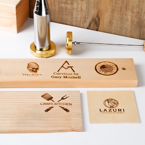 Puede incluir: Una colección de herramientas de marcado en madera y tablas de madera grabadas. Las tablas presentan varios diseños y textos, incluyendo logotipos y los textos "LIAM'S KITCHEN" y "LAZURI - CHICKEN N PIZZA". También se ve un hierro de marcar metálico.