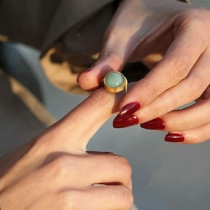Könnte beinhalten: Ein goldener Ring mit einem hellgrünen Edelstein. Der Ring befindet sich an einem Finger einer Person. Die Nägel der Person sind dunkelrot lackiert.