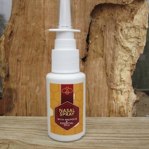 May include: White plastic bottle with a spray nozzle. The bottle has a yellow and brown label with a bee and honeycomb design. The label reads "Nasal Spray with Propolis & Essential Oils".