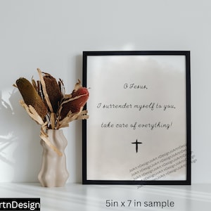 May include: A black framed print with a white background and the text "O Jesus, I surrender myself to you, take care of everything!" in black cursive writing. A small black cross is below the text. The print is sitting on a white surface with a vase of dried flowers in front of it.