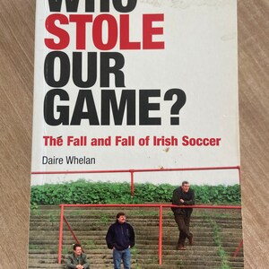 Könnte beinhalten: Ein Buch mit dem Titel "Who Stole Our Game? The Fall and Fall of Irish Soccer" von Daire Whelan. Das Cover zeigt ein Foto von drei Männern, die auf einer Reihe von Betontreppen mit einem roten Geländer stehen.