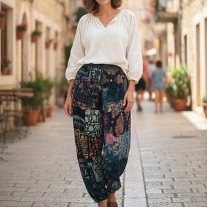 May include: A woman wearing a white long-sleeve blouse with embroidered details and patchwork pants with floral and geometric patterns. The pants are a mix of dark blue, green, and pink. She is wearing brown sandals.