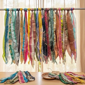 May include: A collection of colourful silk scarves suspended from a metal rail. The scarves, in shades of blue, pink, yellow, and green, are patterned and knotted to the rail. The display creates a vibrant and decorative arrangement.