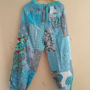 以下が含まれることがあります： ライトブルーのパッチワークパンツ。ウエストはゴムとドローコード仕様。青、緑、グレーの色合いの様々な柄の生地で作られています。木製のハンガーに吊るされています。