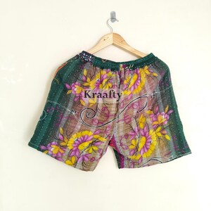 May include: Green, yellow, and purple floral print shorts with the text "Kraafty". The shorts are hanging on a wooden hanger. The shorts have an elastic waistband and are made of a lightweight fabric.