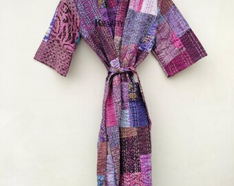Op de afbeelding: Een patchwork badjas in tinten paars, roze en blauw hangt aan een houten hanger. De badjas is gemaakt van verschillende stofvierkanten die met witte draad aan elkaar zijn gestikt. De badjas heeft een strikceintuur en korte mouwen. Het woord "KRAFTY" is zichtbaar.