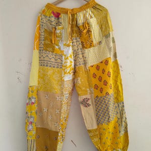 May include: Yellow patchwork pants with a drawstring waist and pockets. The pants are made of various fabrics in shades of yellow, gold, and cream, with floral and geometric patterns. The pants are hanging on a wooden hanger against a white wall.