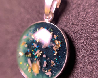 May include: A silver pendant with a round, clear resin charm. The charm contains green, blue, and gold glitter, as well as small white and gold pieces.