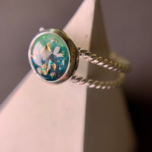 May include: A silver ring with a round, clear resin stone. The stone contains blue, green, and white glitter and gold flakes.