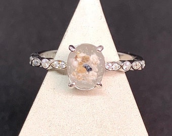May include: A silver ring with a clear oval-shaped stone containing sand and small shells. The band is set with small clear stones.