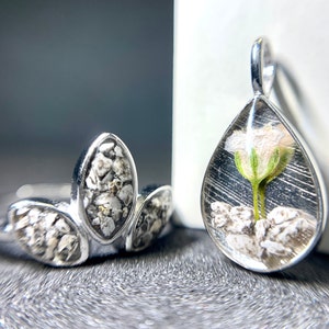 Può includere: Un anello d'argento con tre pietre di forma ovale, ciascuna contenente un piccolo fiore e pietre bianche, e un ciondolo d'argento con una pietra a forma di lacrima che contiene un fiore bianco e pietre bianche.