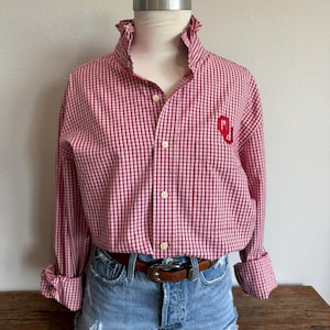 May include: A long-sleeve button-down shirt with a red and white gingham check pattern, featuring a ruffled collar and a red embroidered OU logo on the chest. The shirt is paired with light blue denim shorts and a brown leather belt with a silver buckle.