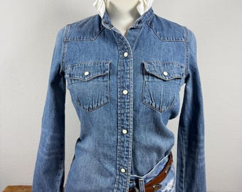 Può includere: Una camicia in denim blu con bottoni, colletto bianco a volant, due tasche sul petto con chiusure a pressione e maniche lunghe. La camicia è infilata in un paio di pantaloncini di jeans azzurri consumati con una cintura in pelle marrone.