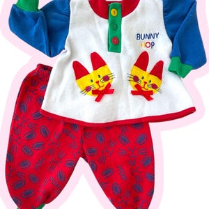 May include: A two-piece baby outfit featuring a white top with blue sleeves and red and yellow bunny face appliqués. The top has the text "BUNNY HOP" and red, green, and yellow accents. The red pants have a bunny pattern.
