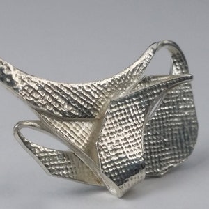 May include: A silver abstract sculpture with a textured surface. The sculpture is made of metal and has a unique, organic shape.