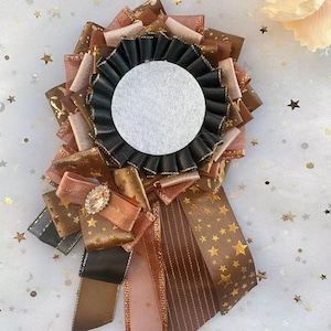 Könnte beinhalten: Eine braune, schwarze und goldene Schleifenrosette mit einem weißen Mittelkreis. Die Rosette hat ein goldenes Sternenmuster und ist mit einem kleinen, klaren Strassstein verziert.