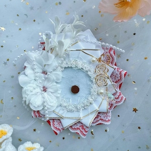 Könnte beinhalten: Ein weiß-rotes Haaraccessoire mit Blumen, Perlen, Spitze und Bändern. Das kreisförmige Design zeigt weiße Stoffblumen, goldfarbene Akzente und einen kleinen braunen Kreis in der Mitte. Der Hintergrund ist weiß mit goldenen Sternen.