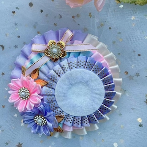 Könnte beinhalten: Eine pastellfarbene Rosette in Lila, Rosa und Weiß mit goldener Einfassung und einem kleinen, weißen, perlmuttartigen Knopf in der Mitte. Die Rosette ist mit einer rosa Blume mit einem weißen, perlmuttartigen Knopf in der Mitte und einer lila Blume mit einem weißen, perlmuttartigen Knopf in der Mitte verziert.