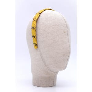 Może przedstawiać: Żółta opaska do włosów z czarnym wzorem wężowej skóry jest prezentowana na głowie manekina. Opaska to cienka opaska, prawdopodobnie wykonana z tkaniny lub podobnego materiału, i jest zakrzywiona, aby pasowała do głowy.