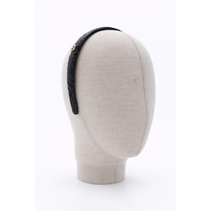 Może przedstawiać: Czarna opaska do włosów o plecionej fakturze z małym, złotym emblematem z boku. Opaska jest prezentowana na głowie manekina na białym tle. Akcesorium do włosów odpowiednie do różnych fryzur.