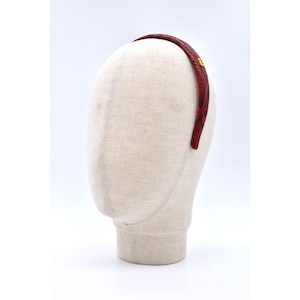 Może przedstawiać: Czerwono-czarna opaska do włosów z wzorem skóry węża i małym złotym emblematem. Opaska jest prezentowana na głowie manekina na białym tle. Stylowy dodatek do włosów.