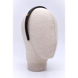 Może przedstawiać: Czarna opaska do włosów o fakturze skóry węża z małym, złotym emblematem. Akcesorium jest prezentowane na głowie manekina. Tło jest białe. Jest przeznaczona do noszenia na głowie.