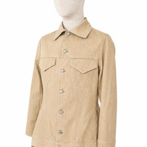 May include: A beige denim jacket with a traditional collar, two chest pockets, and button fastenings. The jacket is displayed on a mannequin. The jacket has long sleeves and a tailored fit. The color is a light tan.