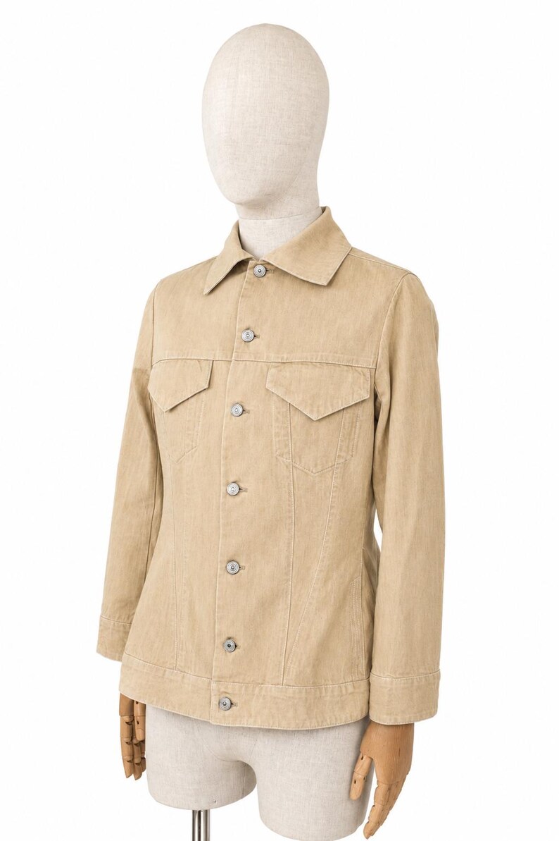 May include: A beige denim jacket with a traditional collar, two chest pockets, and button fastenings. The jacket is displayed on a mannequin. The jacket has long sleeves and a tailored fit. The color is a light tan.