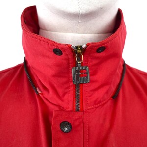 May include: Close-up of a red jacket collar with a black zipper and a logo tag. The jacket has a high collar with black accents and black buttons. The zipper is gold-colored.