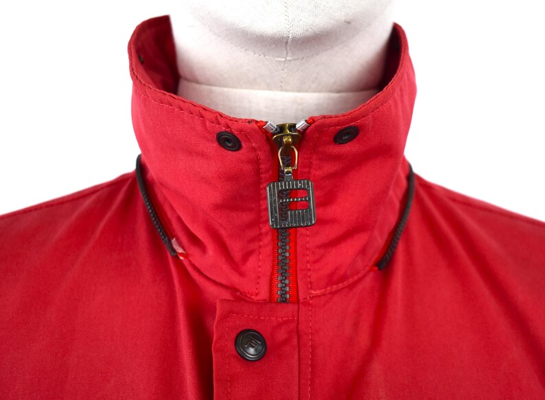 May include: Close-up of a red jacket collar with a black zipper and a logo tag. The jacket has a high collar with black accents and black buttons. The zipper is gold-colored.