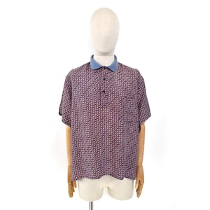 Può includere: Una polo a maniche corte con base blu navy scuro e un motivo geometrico ripetuto di piccole forme rosse e bianche. Il capo presenta un colletto azzurro a tinta unita e un'abbottonatura frontale con due bottoni scuri.