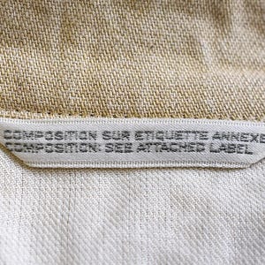 May include: A close-up shot of a garment label, with the text "COMPOSITION SUR ETIQUETTE ANNEXE COMPOSITION: SEE ATTACHED LABEL". The label is white with black text, sewn into the beige and white fabric of the garment.